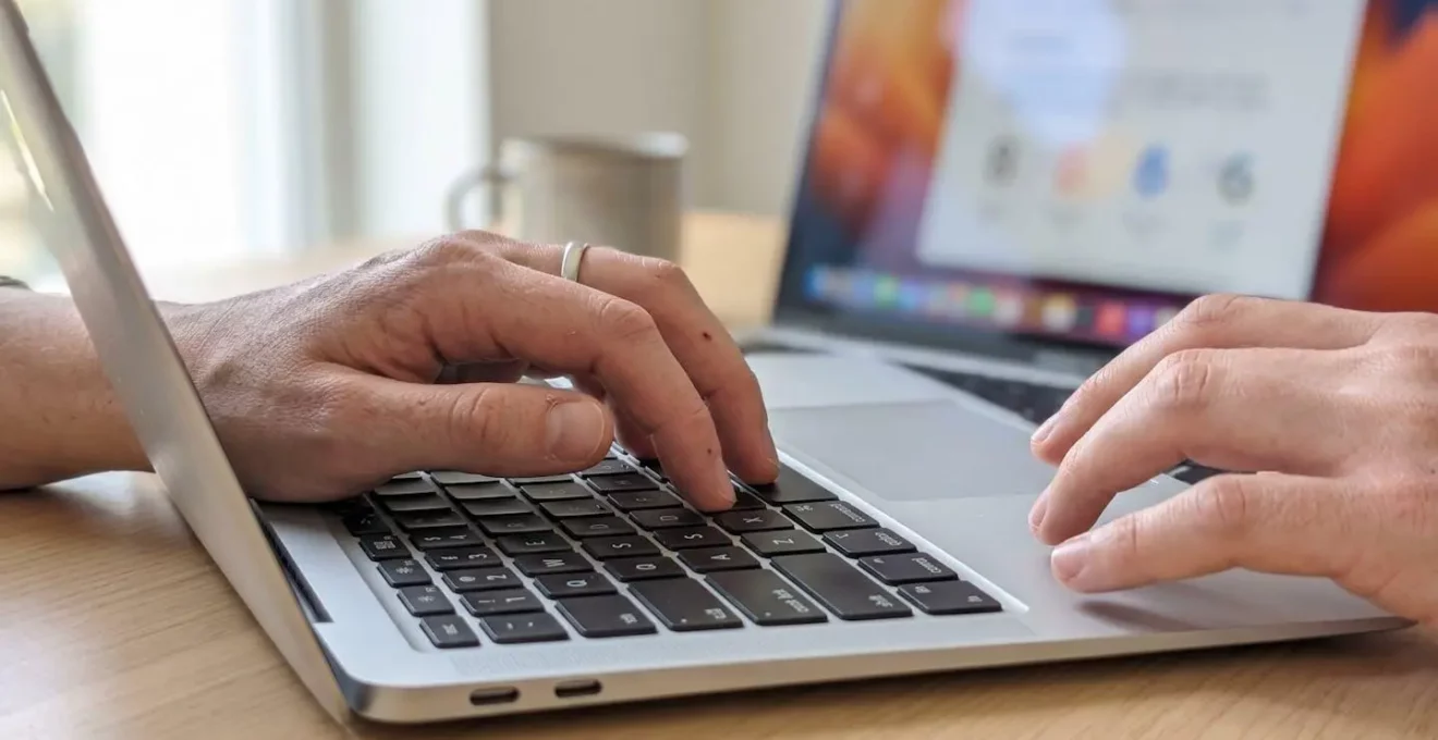 Gros plan sur des mains tapant sur un clavier d'ordinateur portable moderne, avec en arrière-plan flouté un écran affichant des chiffres