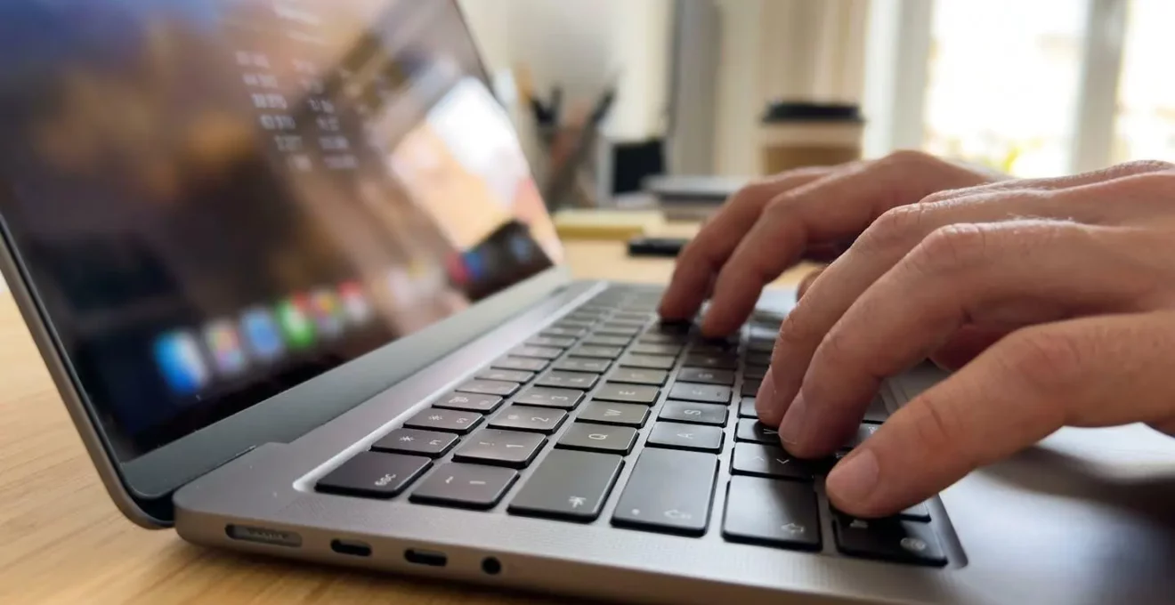 Gros plan sur des mains tapant sur un clavier d'ordinateur portable moderne, avec en arrière-plan flouté un écran affichant des chiffres